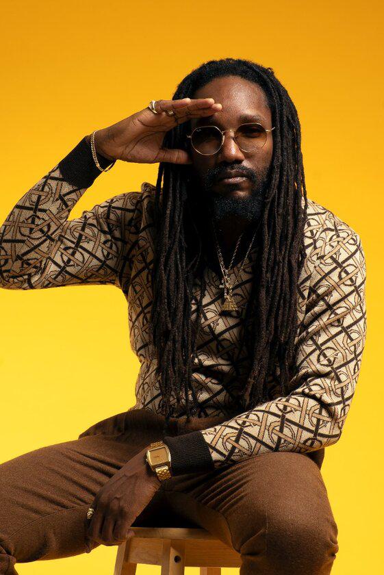 Kabaka Pyramid seated on a stool against a yellow background, shading his eyes with one hand